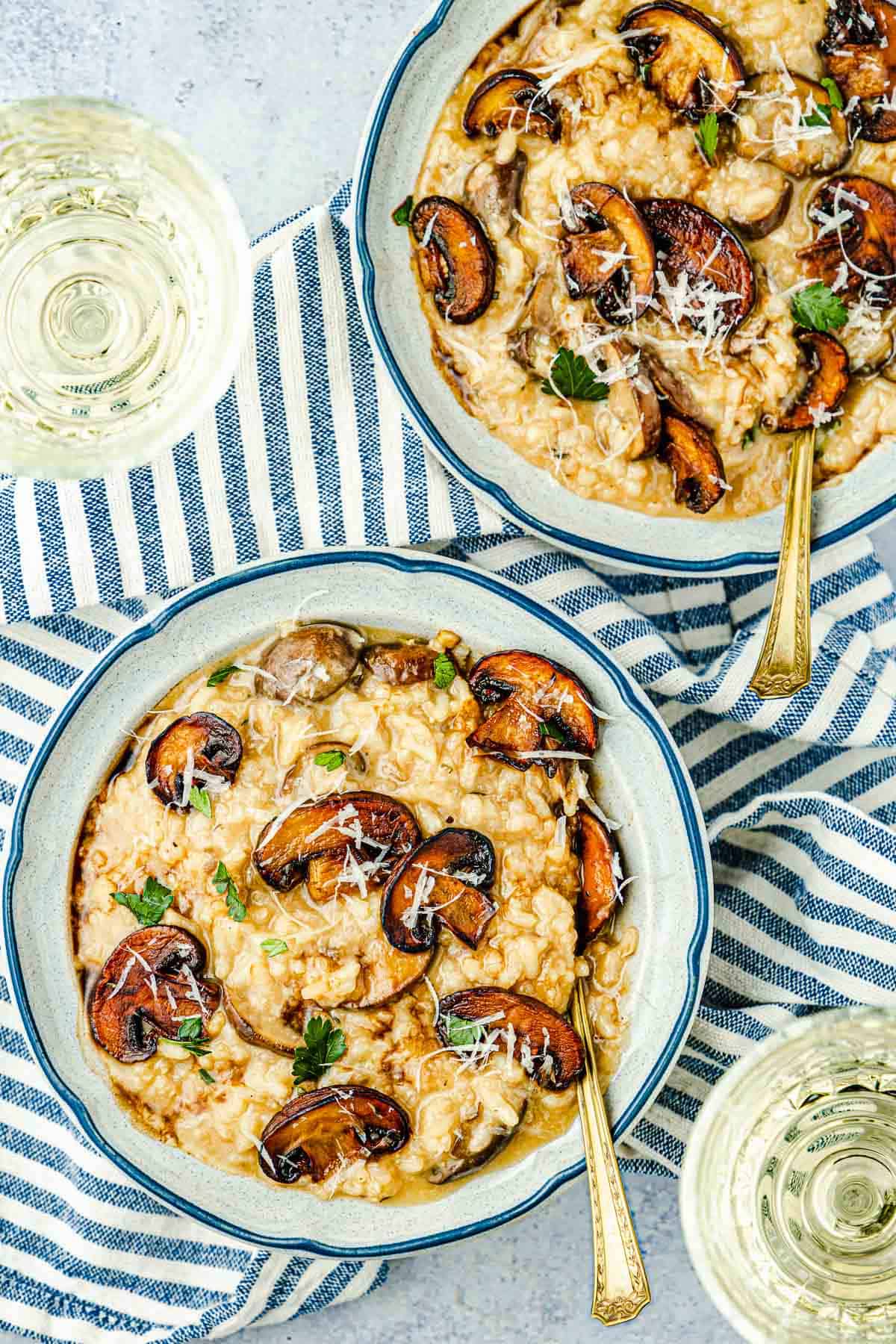 two bowls of mushroom risotto with spoons next to two glasses of white wine.