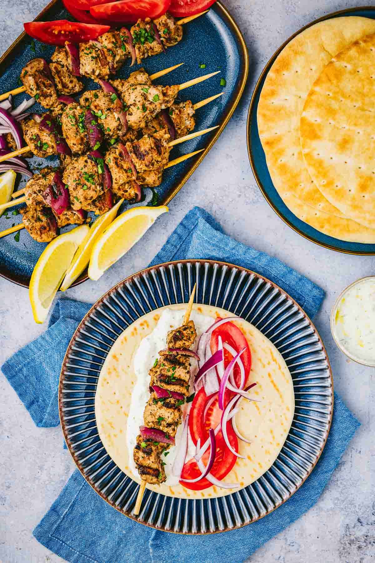 Overhead shot of a pita with tzatziki, red onion, tomato, and a pork souvlaki skewer, with more pork souvlaki skewers, pita bread, and lemon wedges on the side.