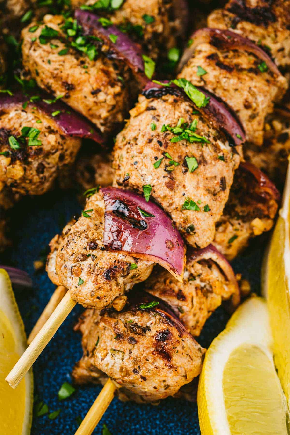 Close up of pork souvlaki skewers with lemon wedges on the side and a sprinkle of chopped fresh parsley.