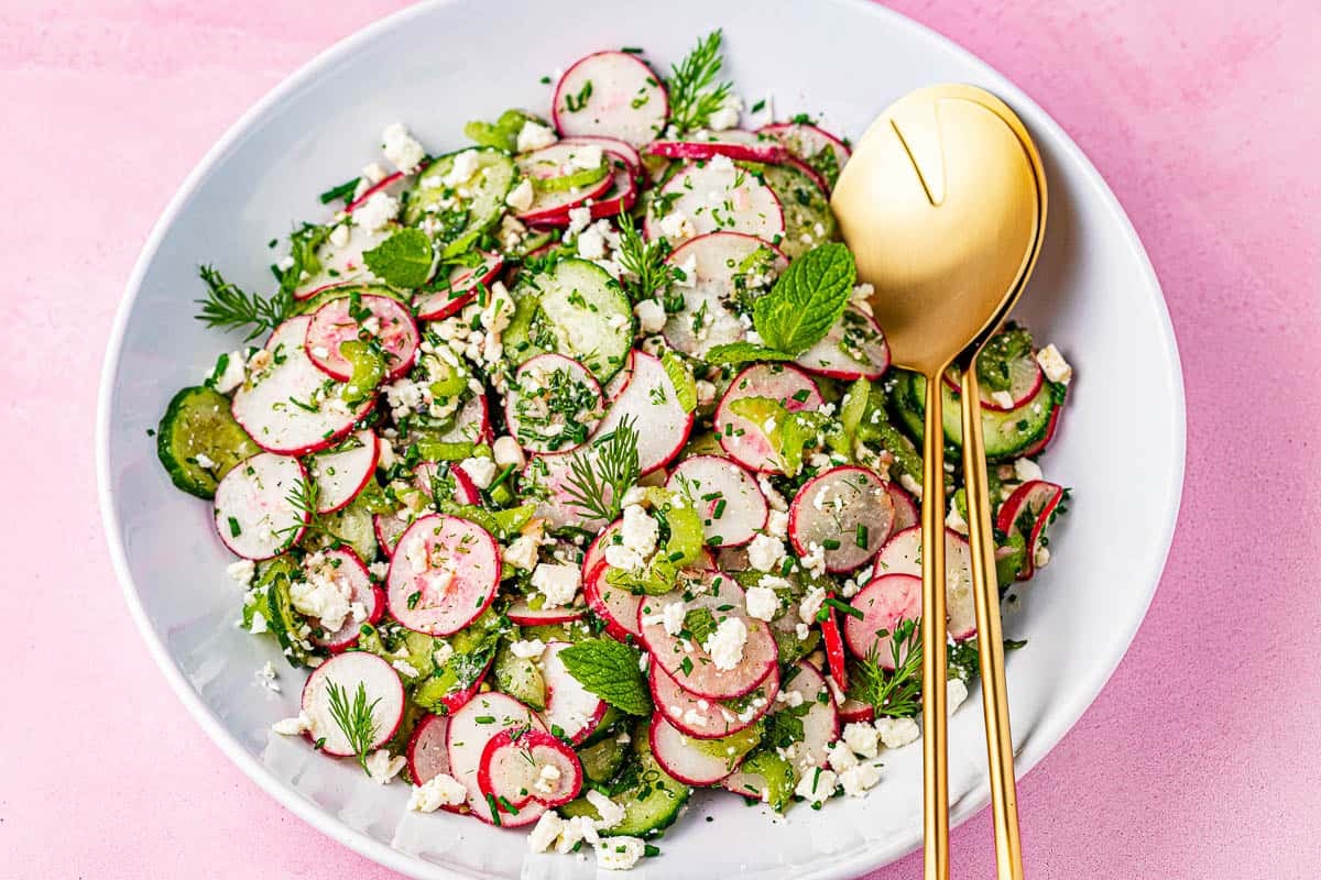cucumber and radish salad in a bowl with gold serving utensils.