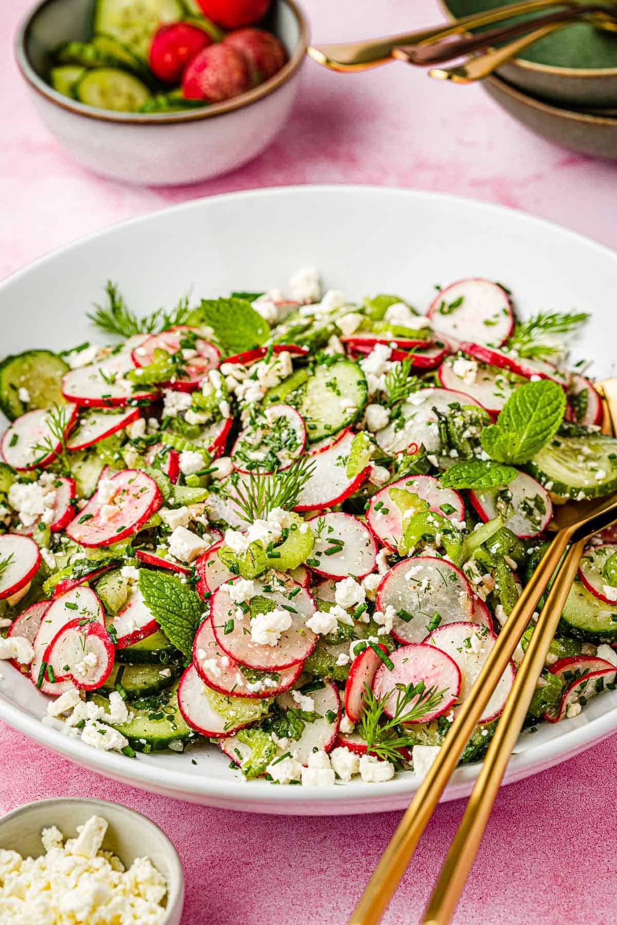 cucumber and radish salad in a bowl with gold serving utensils next to a bowl of radishes and cucumber and a bowl of crumbled goat cheese.