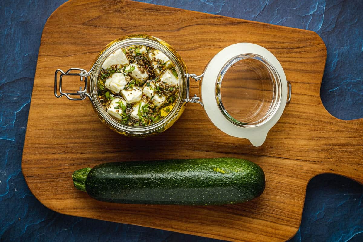 ingredients for zucchini roll ups including marinated feta cubes in a jar and one whole zucchini on a wooden tray.