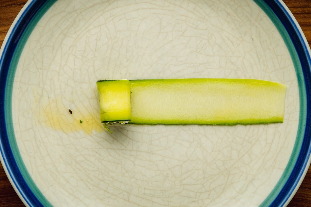 a cube of marinated feta being rolled up in a long thin ribbon of zucchini on a plate.