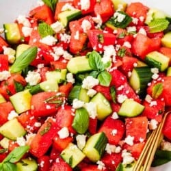 close up of a watermelon salad in a bowl with gold serving utensils.