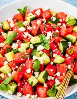 close up of a watermelon salad in a bowl with gold serving utensils.