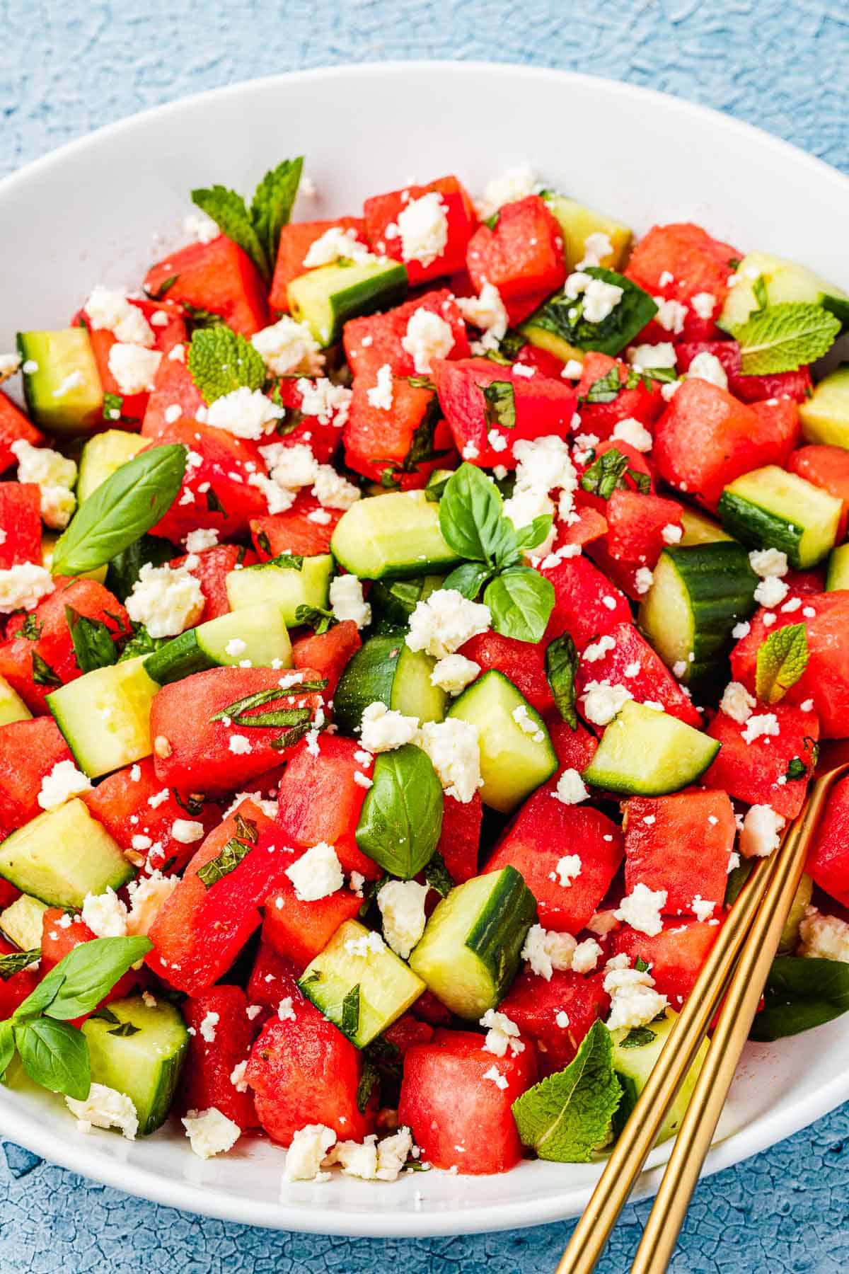 close up of a watermelon salad in a bowl with gold serving utensils.