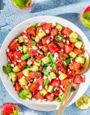 overhead photo of watermelon salad in a bowl with gold serving utensils next to three glasses of water and a bowl of crumbled feta.