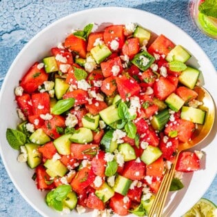 overhead photo of watermelon salad in a bowl with gold serving utensils next to three glasses of water and a bowl of crumbled feta.