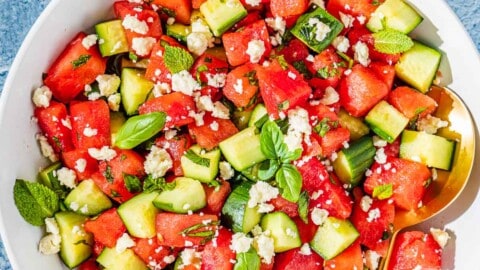 overhead photo of watermelon salad in a bowl with gold serving utensils next to three glasses of water and a bowl of crumbled feta.