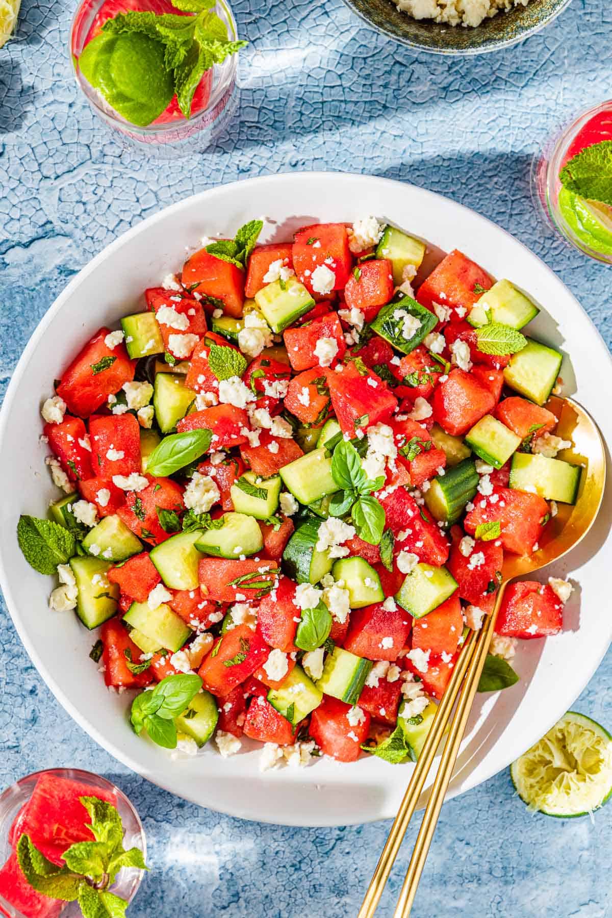 overhead photo of watermelon salad in a bowl with gold serving utensils next to three glasses of water and a bowl of crumbled feta.
