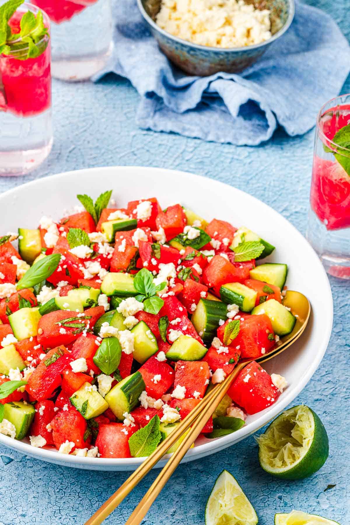 watermelon salad in a bowl with gold serving utensils in front of two glasses of water and a bowl of crumbled feta.