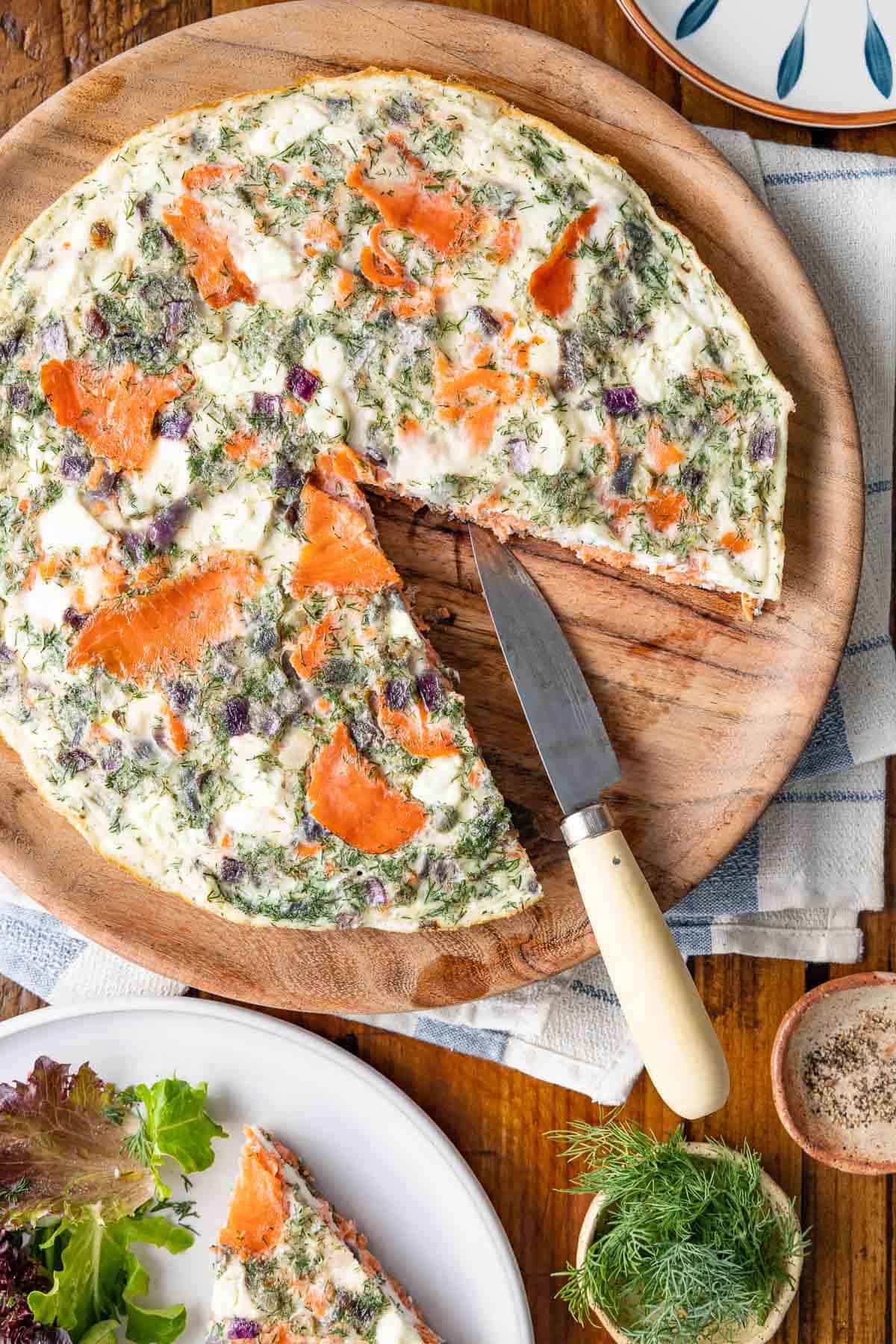 a cooked egg white frittata on a serving tray with a slice missing next to a slice of an egg white frittata on a plate with a small salad and a bowl of fresh dill.