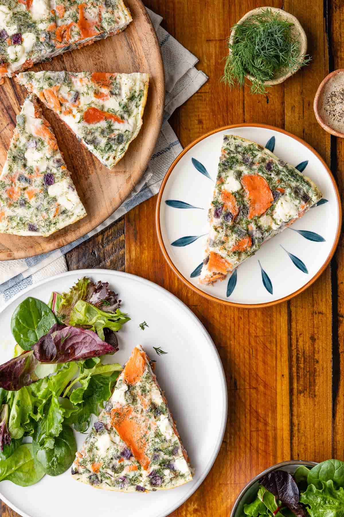 An overhead picture of a slice of an egg white frittata on a plate with a small salad, next to another slice of an egg white frittata on a plate, a sliced up egg white frittata on a serving platter, and a bowl of fresh dill.