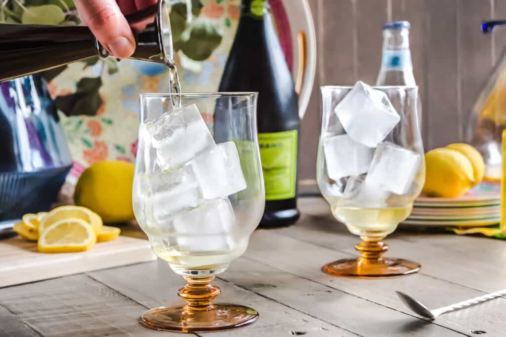 prosecco being poured into a glass filled with ice in front of another ice-filled glass, a bottle of prosecco, a plate of lemons and a cutting board with slices lemons.