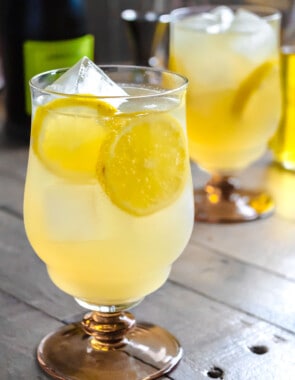 A limoncello spritz garnished with slices of lemon in an ice-filled glass in front of another limoncello spritz and a bottle of prosecco.