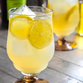 A limoncello spritz garnished with slices of lemon in an ice-filled glass in front of another limoncello spritz and a bottle of prosecco.