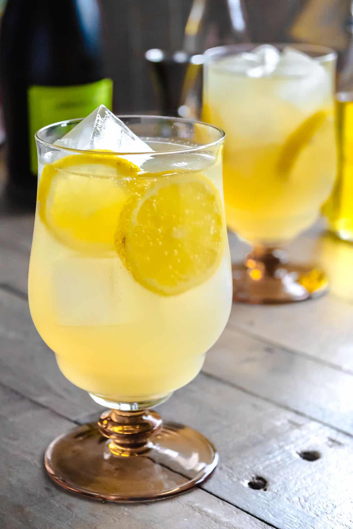 Un spritz au limoncello garni de tranches de citron dans un verre rempli de glace devant un autre spritz au limoncello et une bouteille de prosecco.