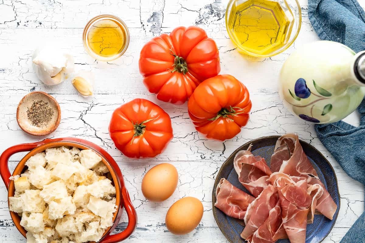 ingredients for salmorejo spanish cold tomato soup including tomatoes, cubes of bread, olive oil, garlic, eggs, pepper, sherry vinegar, and prosciutto.