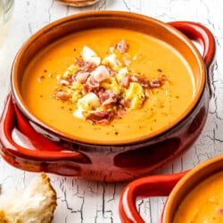 close up of a bowl of salmorejo spanish cold tomato soup topped with chopped hard boiled eggs and prosciutto, next to another bowl of soup, some crusty bread, a glass of wine, and a bowl chopped eggs and prosciutto.