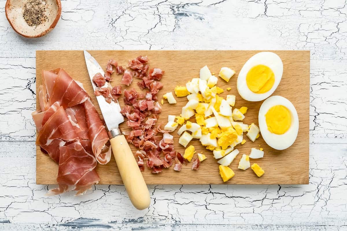a cutting board with prosciutto chopped with a knife, chopped eggs, two hard boiled egg halves next to a bowl of pepper.