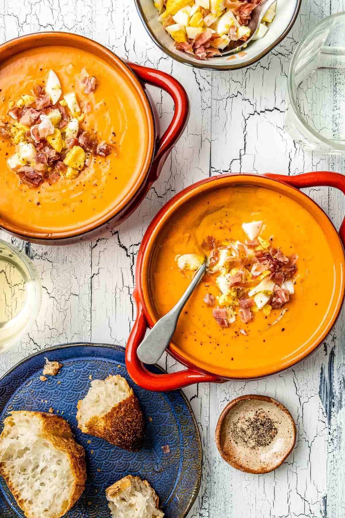 two bowls of salmorejo spanish cold tomato soup topped with chopped hard boiled eggs and prosciutto, next to a plate of crusty bread two beverages, and a bowl of the chopped eggs and prosciutto.