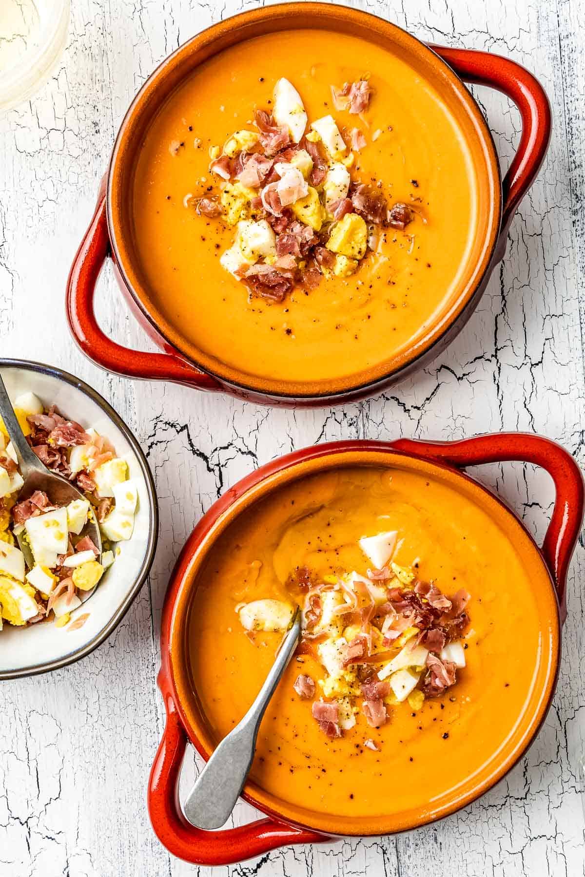 two bowls of salmorejo spanish cold tomato soup topped with chopped hard boiled eggs and prosciutto next to a bowl of chopped eggs and prosciutto.