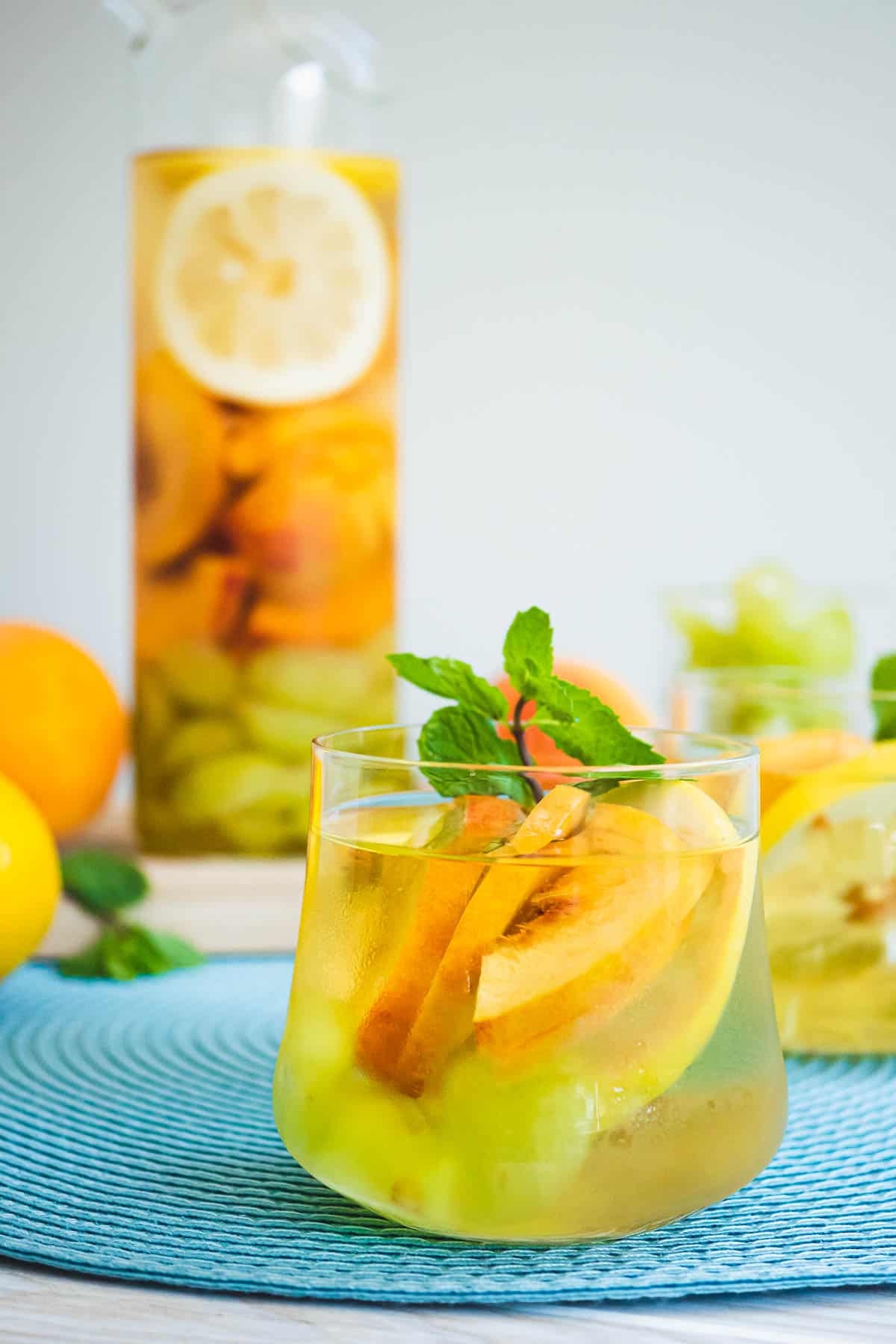 Glass of white wine sangria with sliced peaches, a garnish of fresh mint, and a pitcher with more sangria in the background.