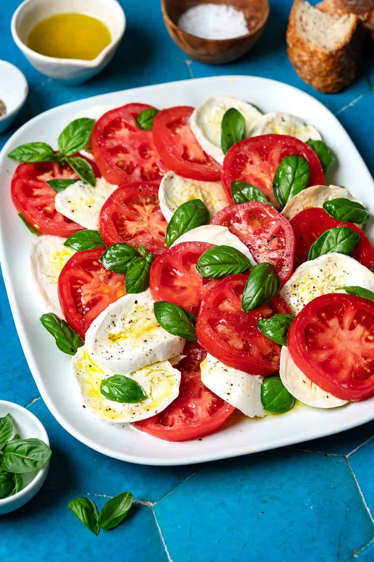 a caprese salad on a platter surrounded by pieces of crusty bread and bowls of olive oil, salt and basil leaves.