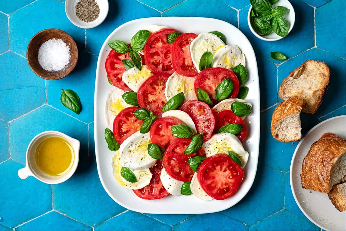 overhead photo of a caprese salad on a platter surrounded by pieces of crusty bread and bowls of olive oil, salt, pepper and basil leaves.