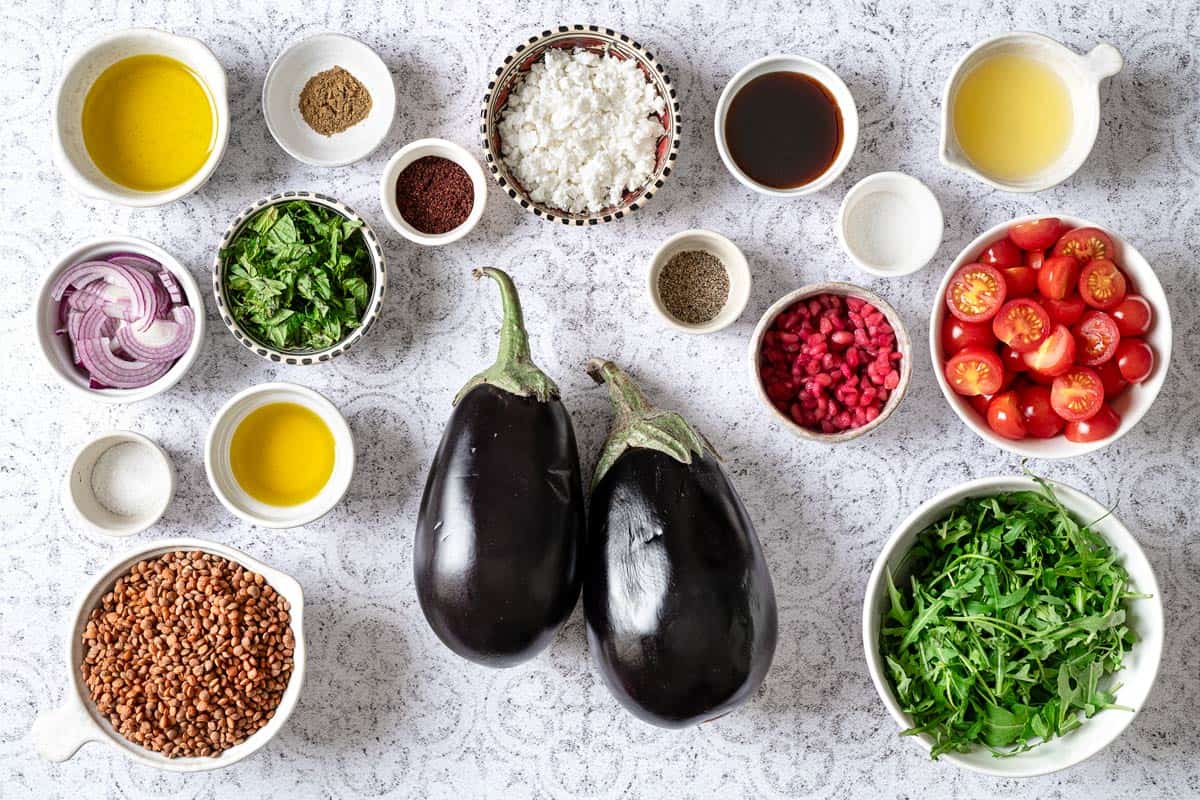 Ingredients for eggplant salad, including cherry tomatoes, sumac, lemon, salt, pepper, lentils, olive oil, feta cheese, red onion, arugula, cumin, and mint.