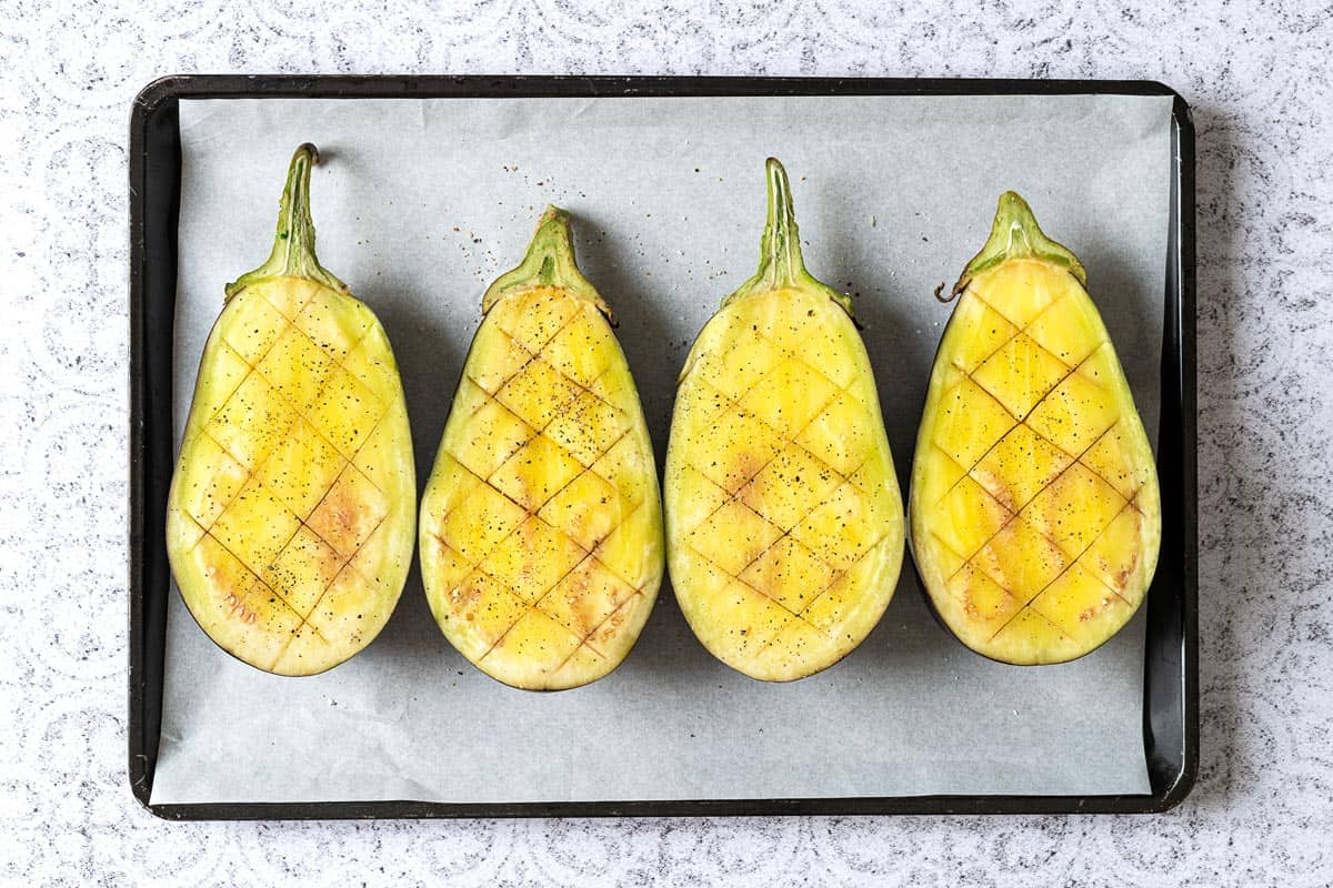 Four raw eggplant halves on a sheet tray with a diamond pattern slices.