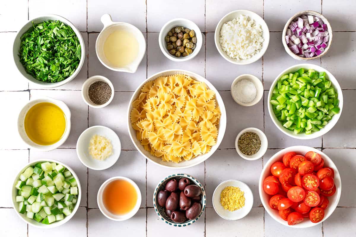 ingredients for greek pasta salad including parsley, lemon juice, lemon zest, capers, feta, red onion, olive oil, pepper, oregano, bell pepper, salt, cucumber, cherry tomatoes, kalamata olives and farfalle pasta.