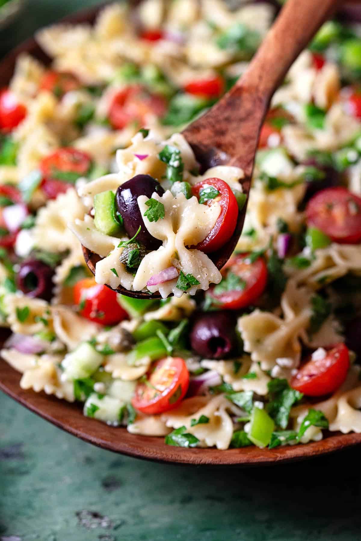 a close up of a bite of greek pasta salad on a wooden serving spoon lifted over an entire greek pasta salad in a serving bowl.