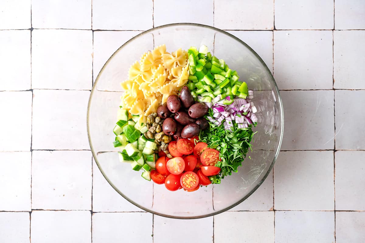 farfalle pasta, diced cucumber, chopped bell pepper, kalamata olives, chopped red onion, chopped parsley, halved cherry tomatoes and capers in a bowl, prior to mixing.