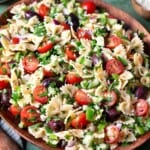 a greek pasta salad in a serving bowl next to a bowl of chopped parsley, a bowl of kalamata olives, and a bowl of crumbled feta cheese.