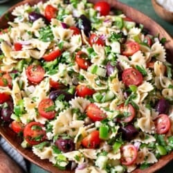 a greek pasta salad in a serving bowl next to a bowl of chopped parsley, a bowl of kalamata olives, and a bowl of crumbled feta cheese.