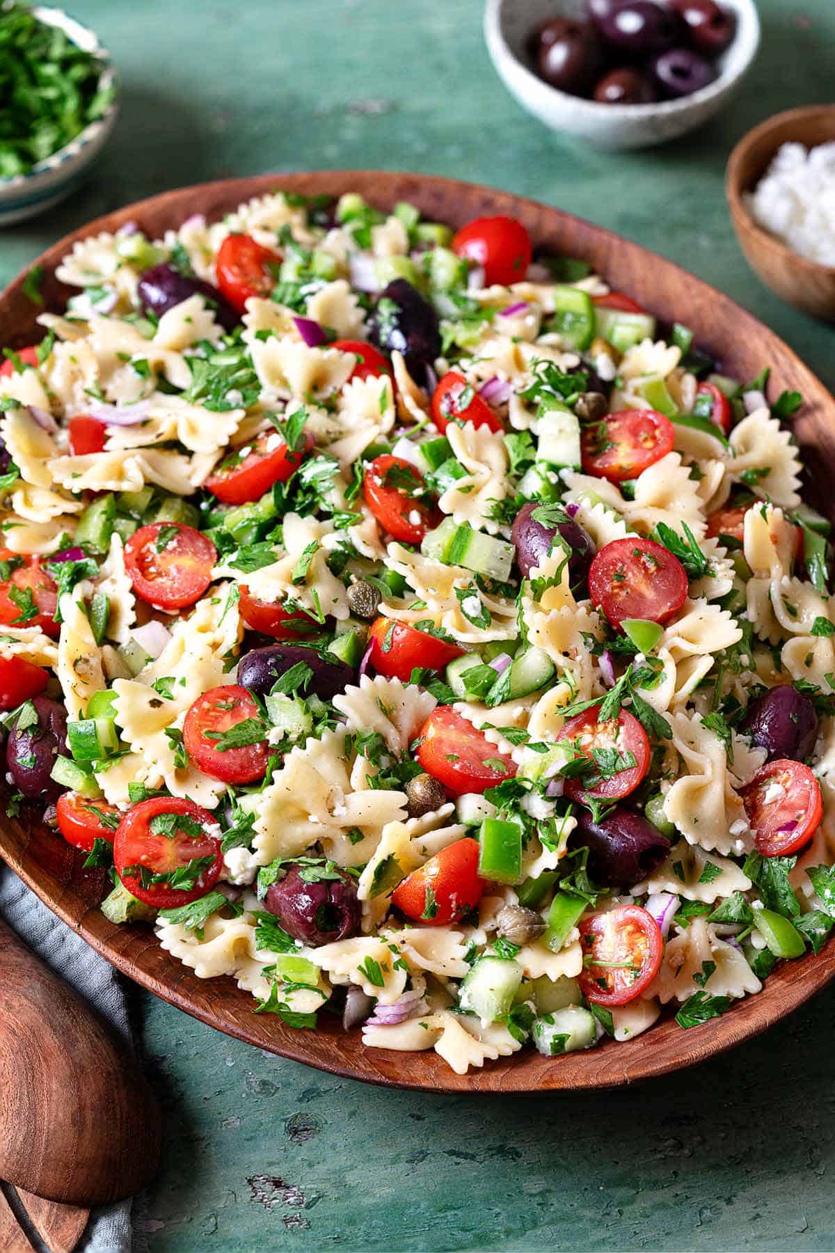 a greek pasta salad in a serving bowl next to a bowl of chopped parsley, a bowl of kalamata olives, and a bowl of crumbled feta cheese.