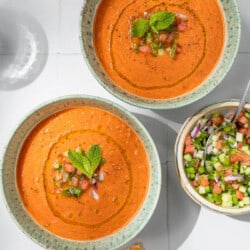 Overhead photo of 2 bowls of watermelon gazpacho topped with, diced watermelon, cucumber, green pepper, olive oil, black pepper and mint next to a bowl of these garnishes and two spoons.