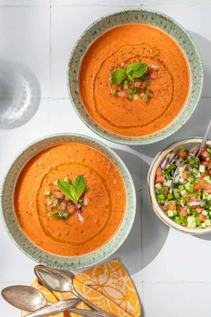 Overhead photo of 2 bowls of watermelon gazpacho topped with, diced watermelon, cucumber, green pepper, olive oil, black pepper and mint next to a bowl of these garnishes and two spoons.