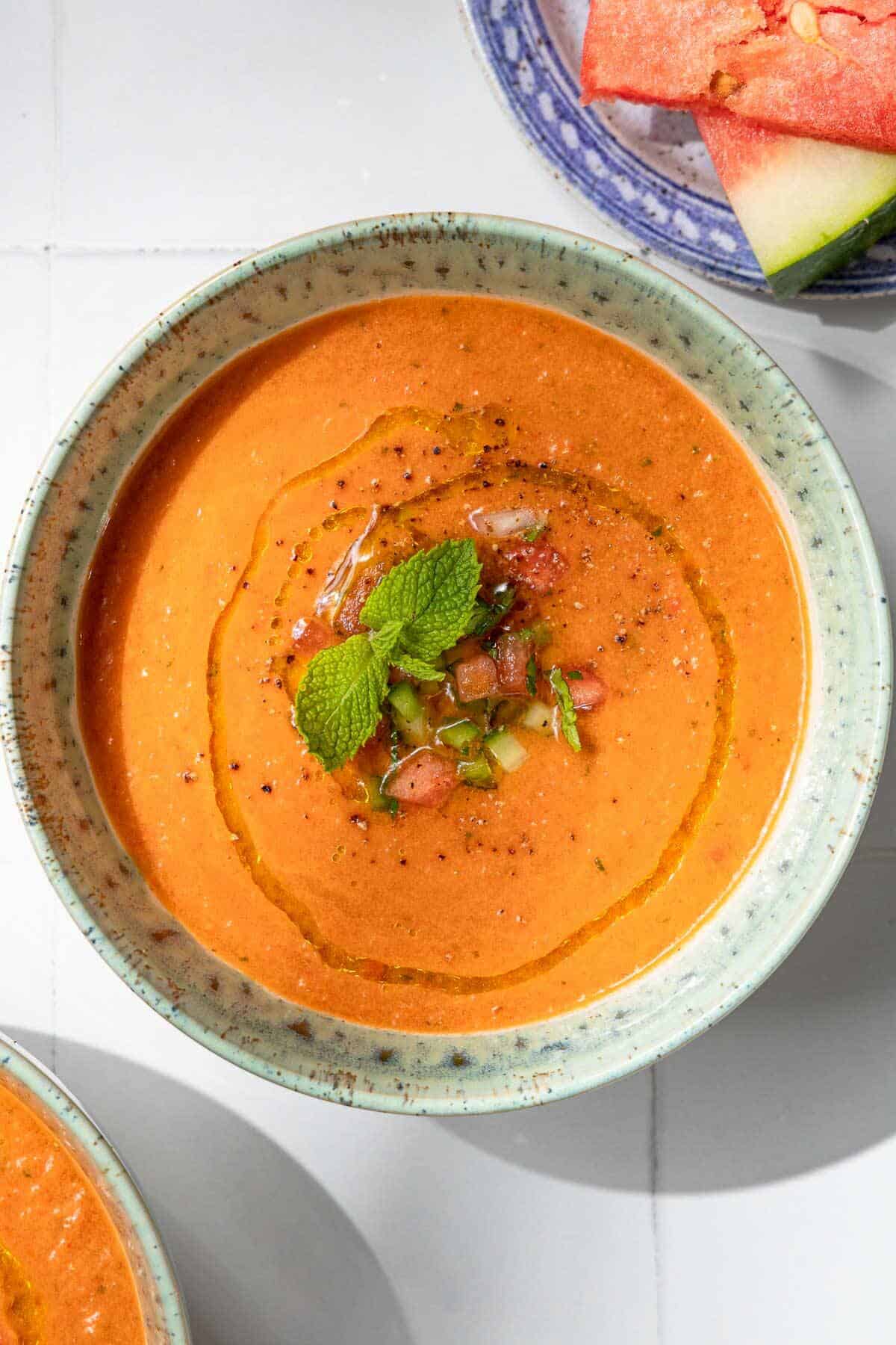 Overhead photo of a bowl of watermelon gazpacho topped with, diced watermelon, cucumber, green pepper, olive oil, black pepper and mint.