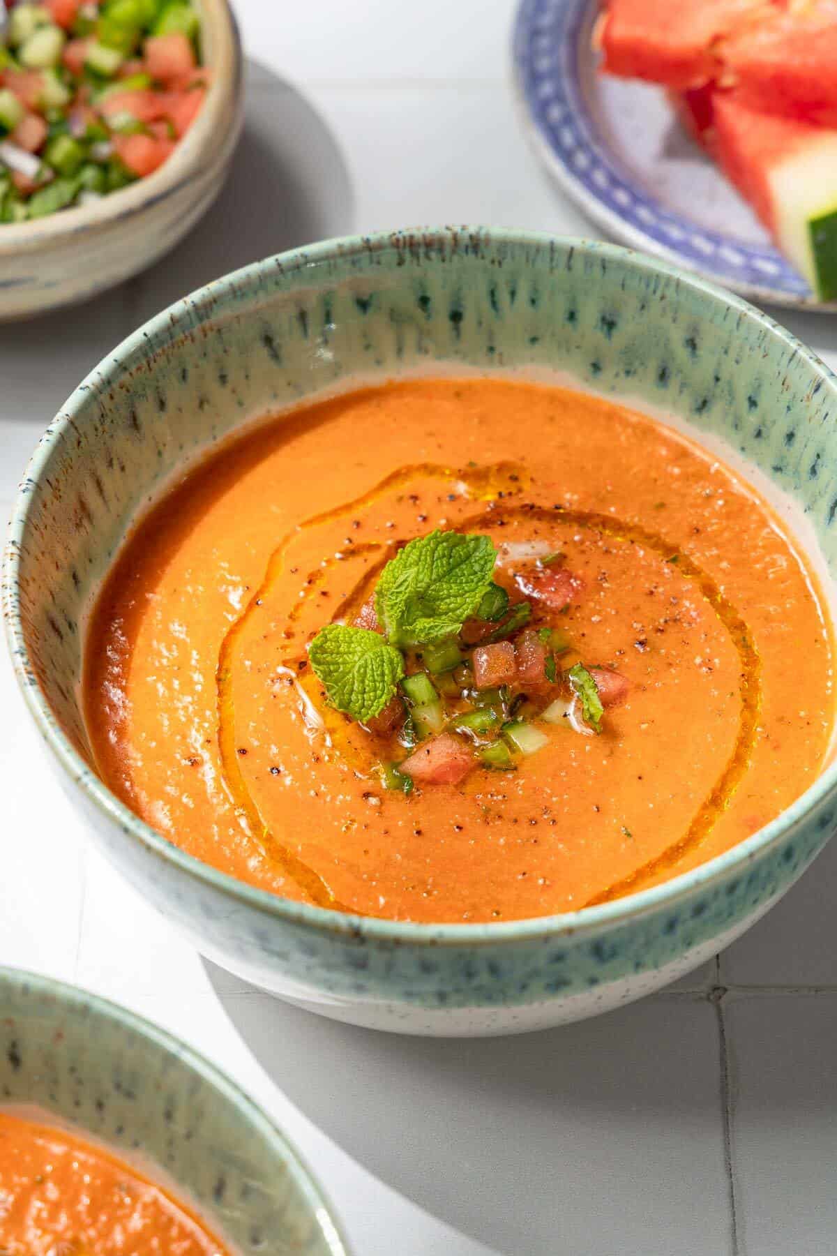 a close up of a bowl of watermelon gazpacho topped with, diced watermelon, cucumber, green pepper, olive oil, black pepper and mint.