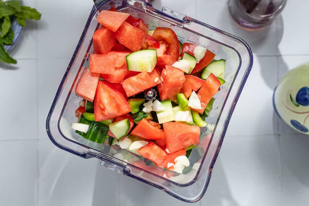 overhead photo of chopped watermelon, cucumber, tomatoes, garlic, red pepper and green pepper in the pitcher of a blender.