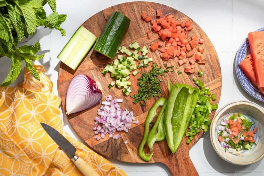 diced tomatoes, mint, cucumber, green onion and red onion on a cutting board along with 2 cucumber halves, a quarter of red onion and large pieces of green bell pepper.