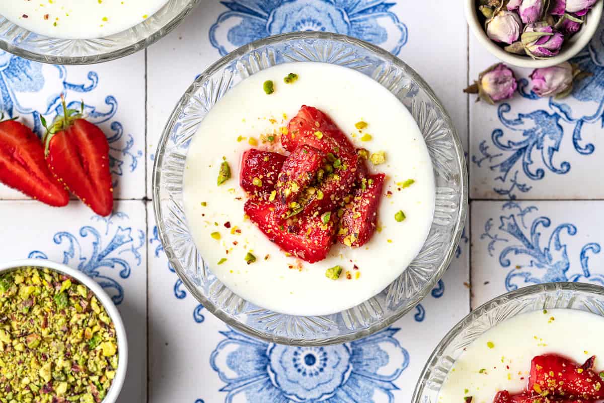 Overhead photo of 3 bowls of Rose Mahalabia (Milk Pudding) topped with strawberries and pistachios next to a bowl of pistachios and a bowl of dried roses.