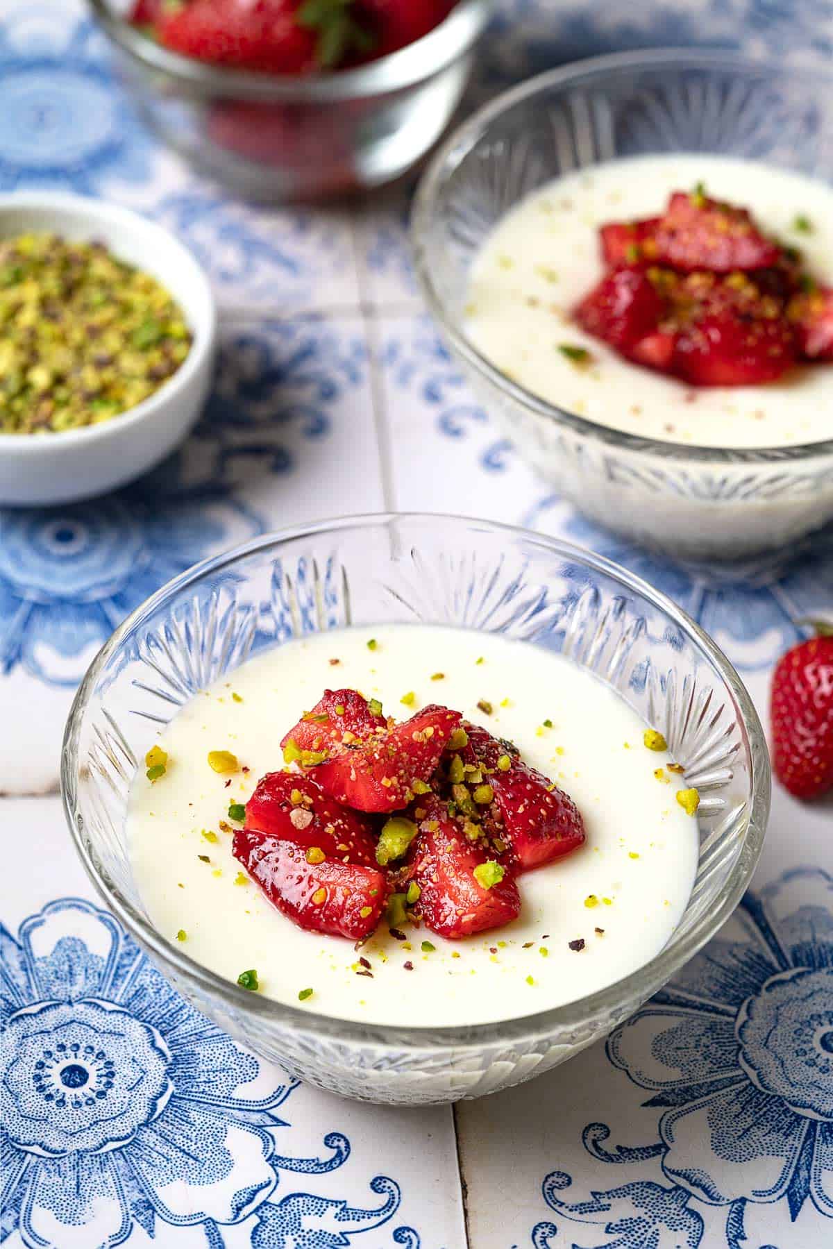 Rose Mahalabia (Milk Pudding) in a bowl topped with strawberries and pistachios in front of another bowl of mahalabia, a bowl of strawberries and a bowl of pistachios.