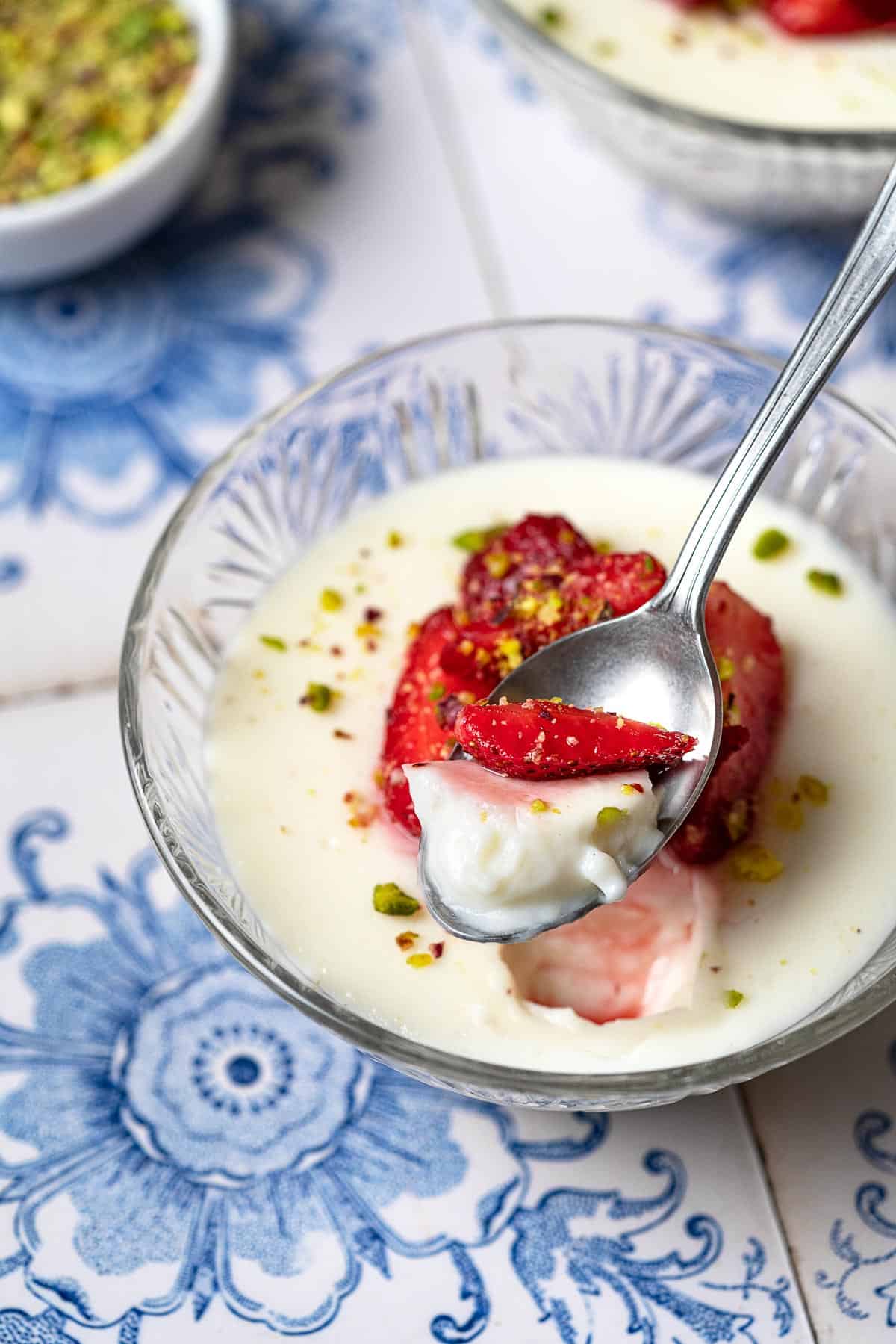 a bite of Rose Mahalabia (Milk Pudding) topped with strawberries and pistachios being lifted out of a bowl with a spoon.