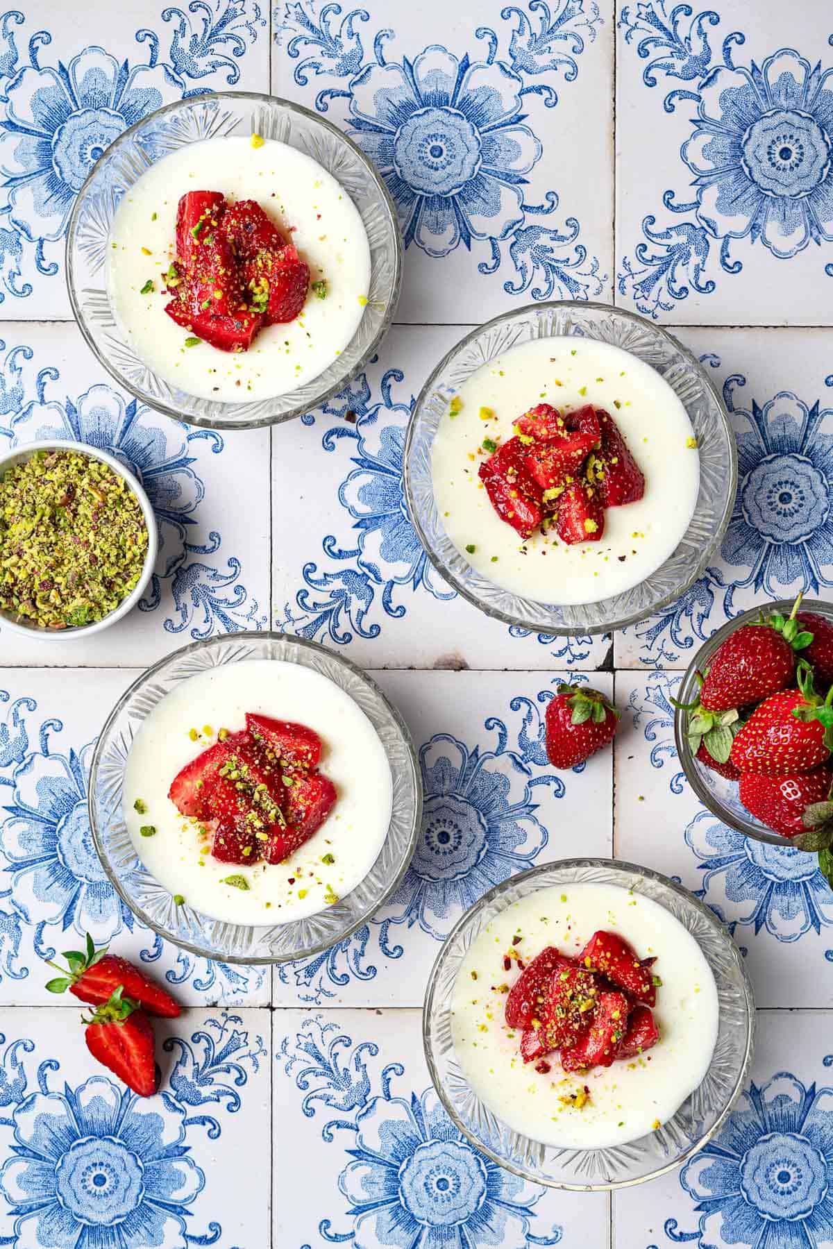 Overhead photo of 4 bowls of Rose Mahalabia (Milk Pudding) topped with strawberries and pistachios next to a bowl of pistachios and a bowl of strawberries.