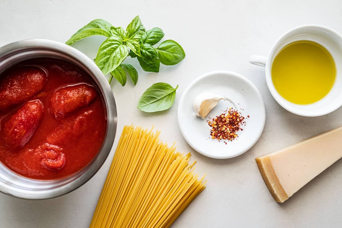 ingredients for pasta pomodoro including whole peeled tomatoes, basil, spaghetti, garlic, red pepper flakes, olive oil and parmesan cheese.