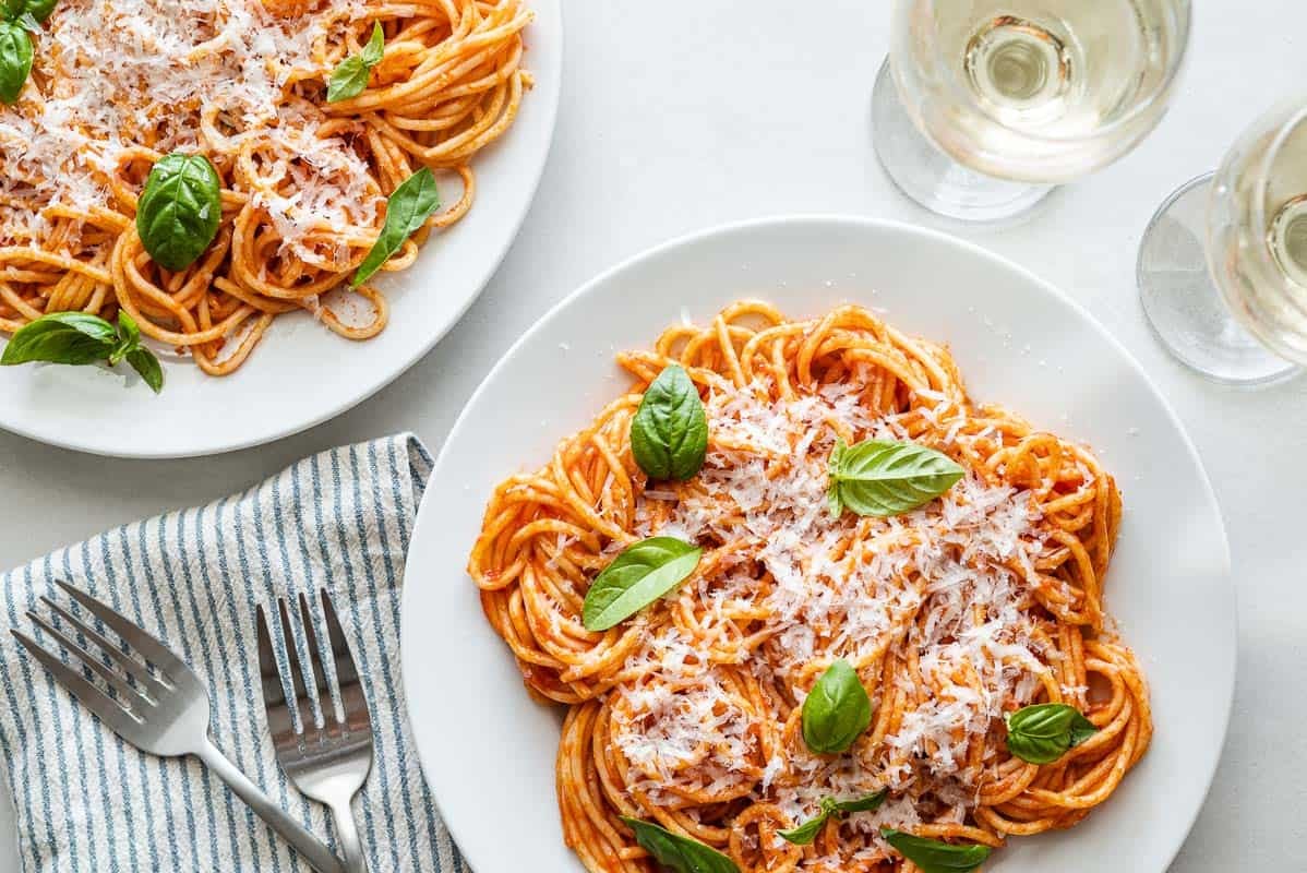 two plates of pasta pomodoro topped with grated parmesan and basil next to two forks, a napkin and two glasses of white wine.