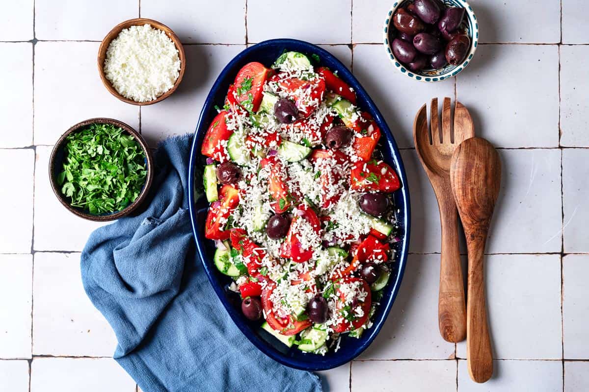 Overhead shot of shopska salad with a sprinkling of white cheese in a large serving platter with wooden serving spoons, more herbs, and olives on the side.